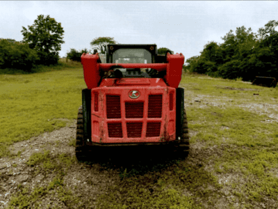 Red skid steer loader with enclosed cab on grassy field, overcast sky.