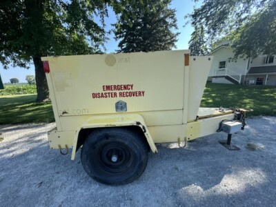 Yellow emergency disaster recovery trailer with single axle and black tire, parked on gravel with trees and houses in the背景.