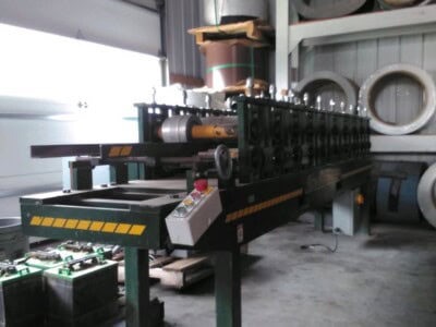 Industrial pipe bending machine in warehouse with rolls of fascia in background.