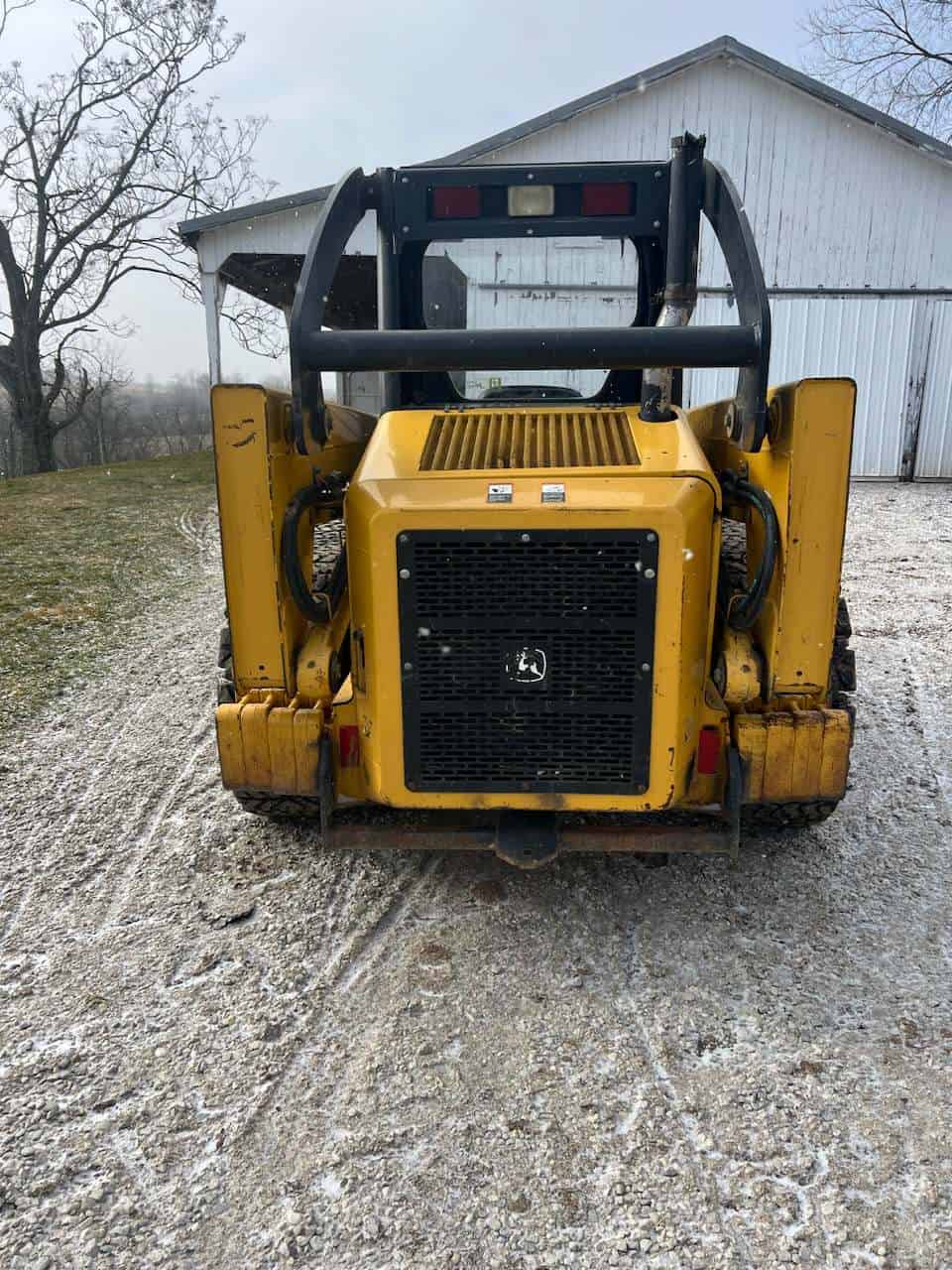 image000001 (21) Yellow compact skid steer loader with protective cage and black grille, parked outdoors near a white barn.