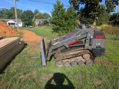 Compact track loader with bucket attachment on grassy lot, residential neighborhood in background.