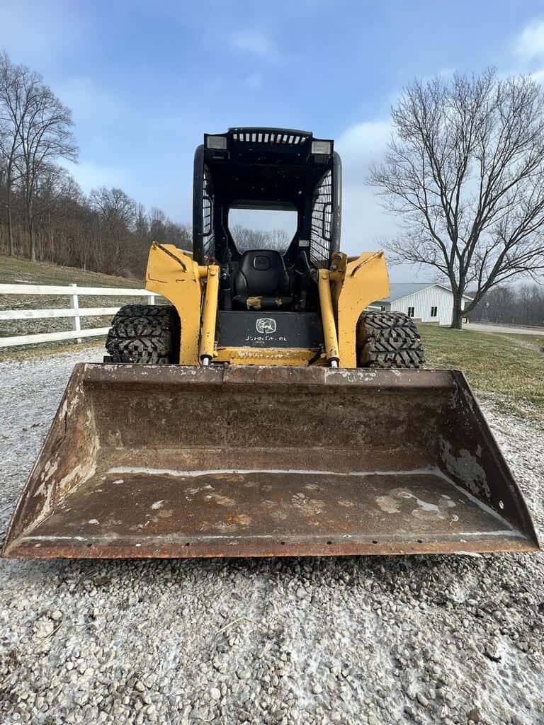 image000002 (18) Yellow skid steer loader with large front bucket on gravel surface, trees and blue sky in background.
