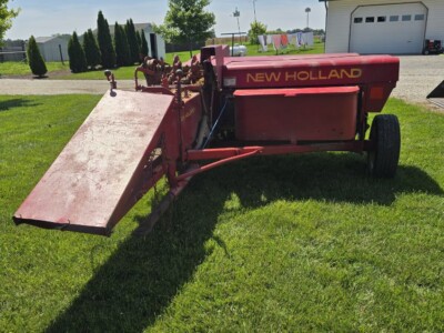 New Holland 273 hay rake with red frame and large wheels on green grass.