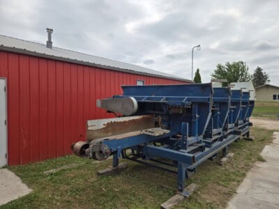 Blue industrial conveyor belt system with rusted rollers outside red building.