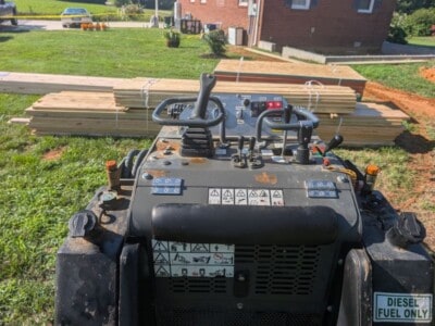 Construction equipment operator's view with control panel and stacks of lumber in background.