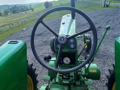 View from the tractor seat showing steering wheel and dashboard of a John Deere 5055E, green with yellow accents, on a farm.