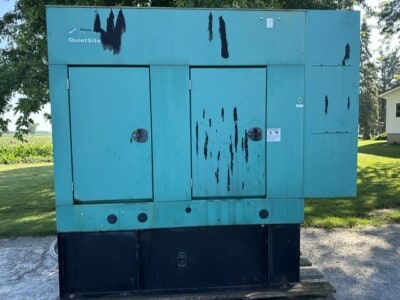 Blue industrial generator with visible rust and wear, outdoors on a concrete pad.