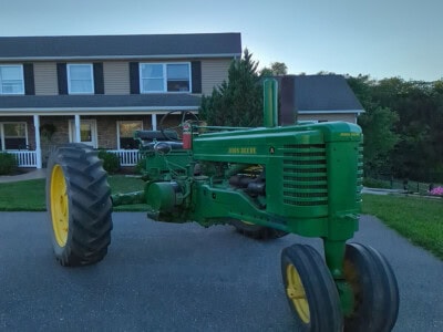 Green vintage John Deere tractor with yellow wheels parked on driveway near house.