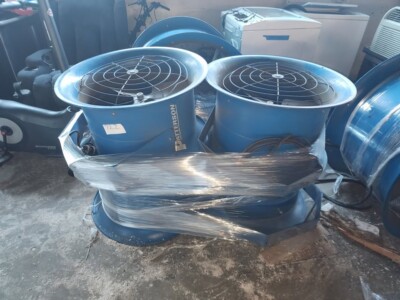 Two blue industrial air circulators with protective grilles, wrapped in plastic, on a concrete floor.