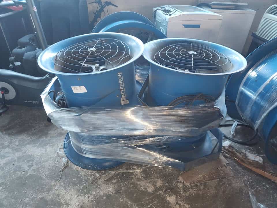 Two blue industrial air circulators with protective grilles, wrapped in plastic, on a concrete floor.
