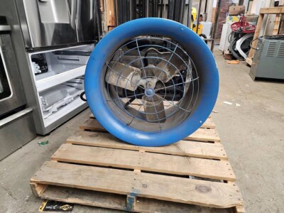Large industrial fan with blue protective frame and metal blades on wooden pallet in warehouse.