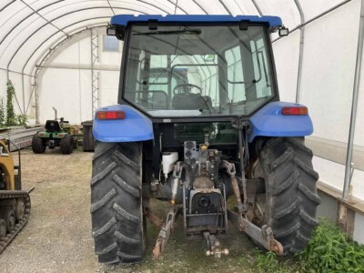 Blue New Holland tractor with enclosed cab and large rear tires in a greenhouse setting.
