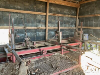 Red logging equipment with multiple saw blades and hydraulic arms in a wooden shed.