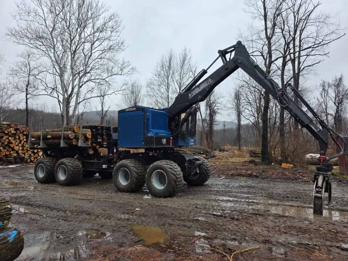 Forestry grapple loader with log pile in muddy outdoor setting.