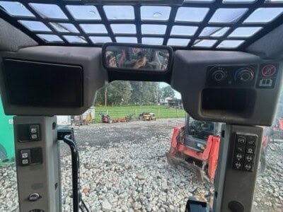 Cabin interior of a 2024 Yanmar TL100VS tractor with control panels and view of the outdoor worksite.