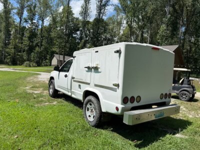 White utility service truck with enclosed cargo area and rear bumper, parked on grassy field with trees in background.
