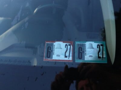 Two vehicle inspection stickers on the lower corner of a car windshield, one red and one blue, with the numbers 6 and 27.
