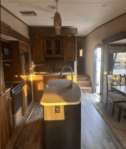 RV kitchen with wooden cabinets, island with sink, and dining area with large window.