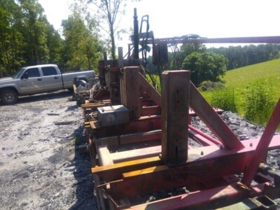 Red circular sawmill with metal framework and cutting blade, outdoors on gravel with trees in background.