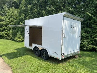 ATC enclosed aluminum concession trailer with side awning and dual axles, parked on grass near trees.