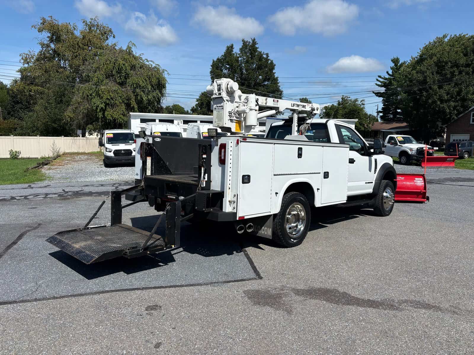 10946 - 2018 Ford F-550 Chassis - 009 White utility truck with bucket lift and equipment storage, parked on asphalt with trees and houses in background.