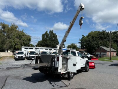 White service truck with crane and bucket, parked on asphalt lot with trees and houses in background.