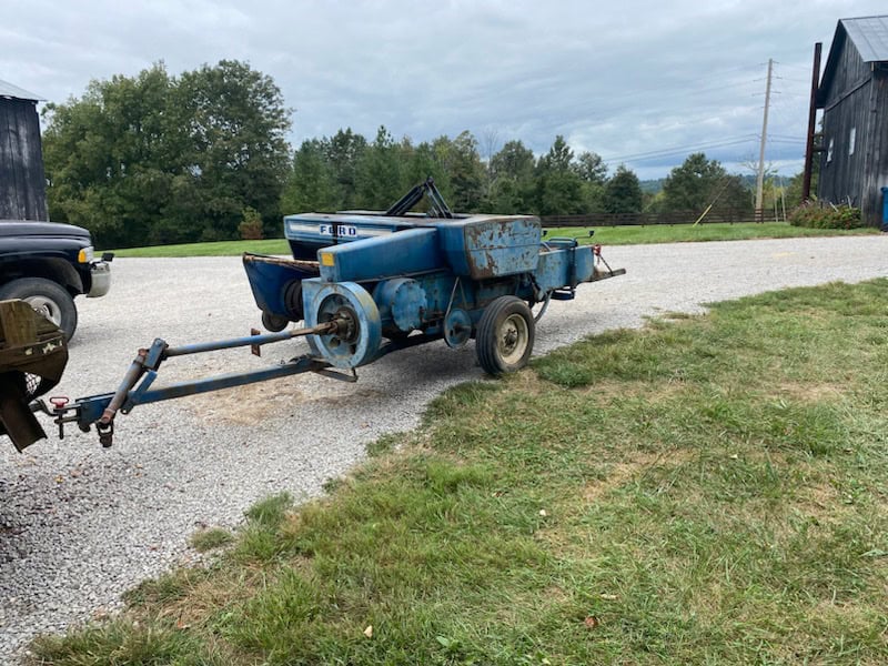 Old blue agricultural seeder machine with rust and wear, on a gravel surface with trees and buildings in background.