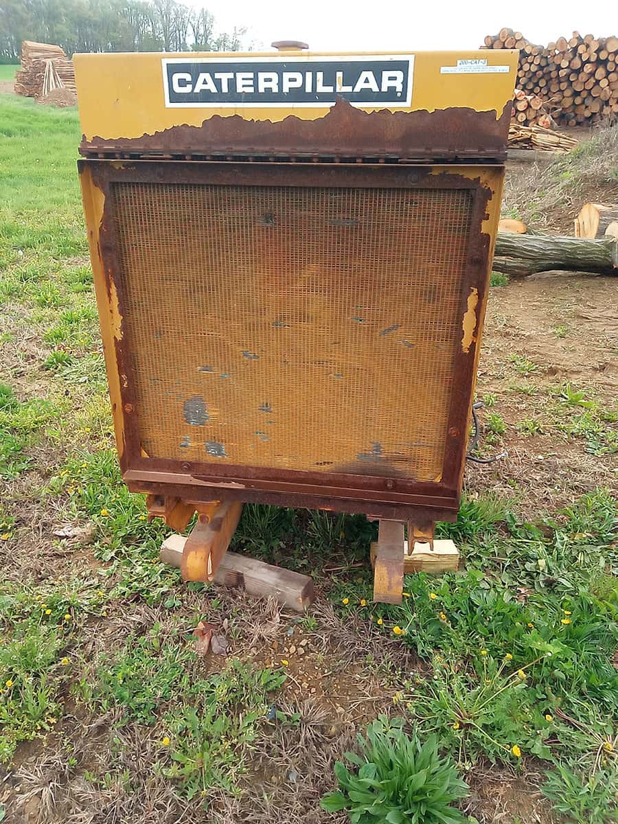 IMG_20260420_092028_628 1 Caterpillar radiator core with rust and wear, outdoor setting with logs and grass.