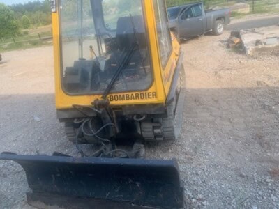 Yellow Bombardier compact excavator with blade attachment on gravel surface.