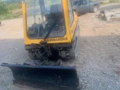 Yellow Bombardier mini excavator with front blade and enclosed cab on gravel surface.