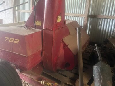 Red New Holland 782 hay baler with large pickup and hydraulic components inside a barn.