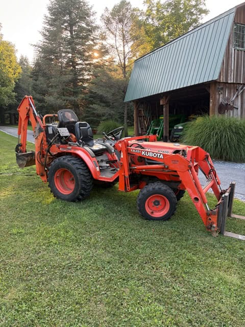 Kubota L Series compact tractor with backhoe attachment, orange color, in a grassy yard with barn and trees.