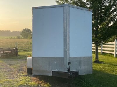 White enclosed utility trailer with tandem axles and ramp door in a rural setting.