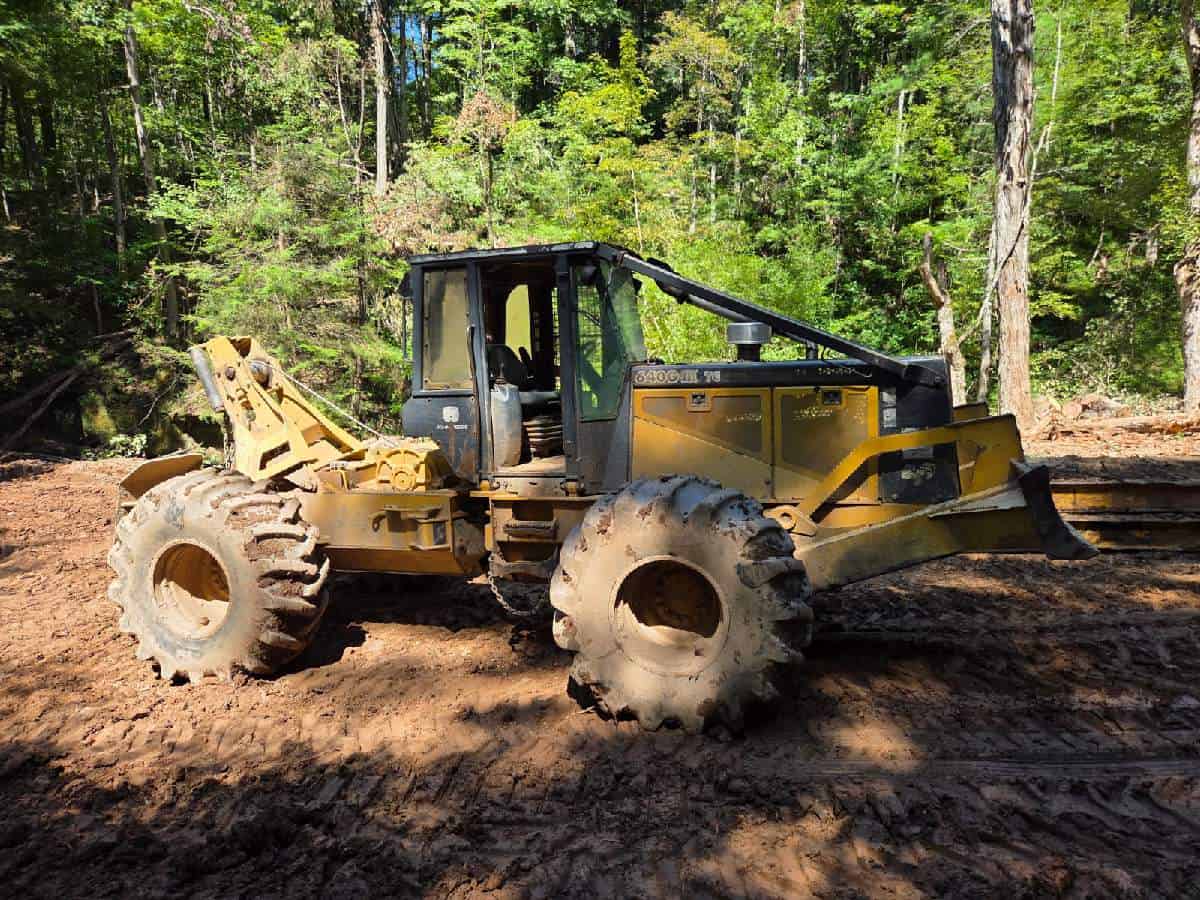 Yellow Caterpillar skid steer loader with enclosed cab on a dirt site surrounded by trees.