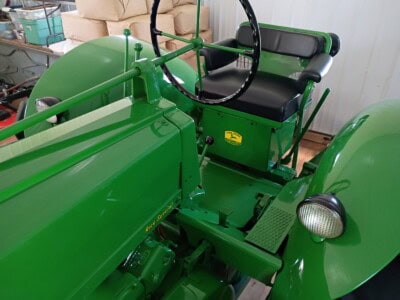 Green vintage John Deere tractor with black seat and steering wheel, indoor setting.