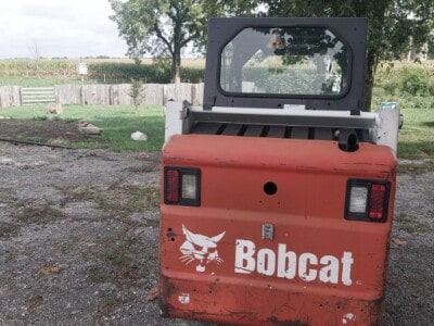 Red Bobcat skid steer loader with black cab and white logo, outdoor setting with trees and fence.