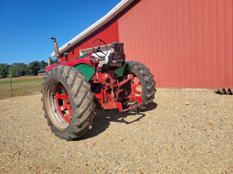 image000003 (7) Red and green vintage tractor with large rear tires and open cab, parked on gravel near a red barn.