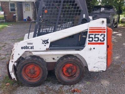 Bobcat 553 skid steer loader with enclosed cab and red wheels, parked outdoors.