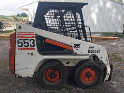 Bobcat 553 skid steer loader with enclosed cab and orange wheels, parked outdoors.