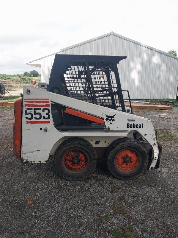 Bobcat 553 skid steer loader with enclosed cab and orange wheels, parked outdoors.