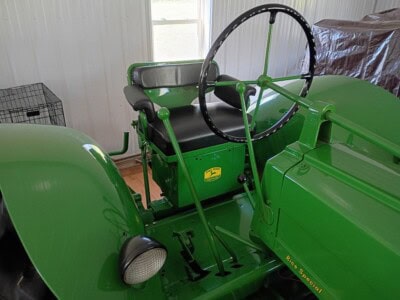 Green vintage tractor with black seat and steering wheel inside a shed.