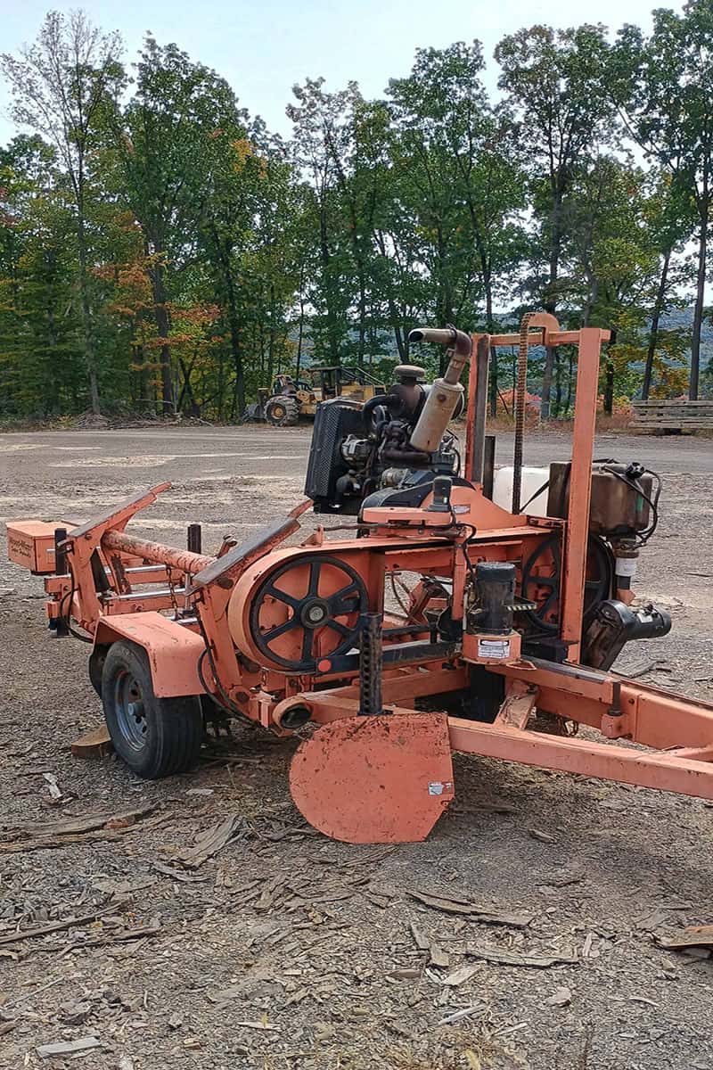 1000010690 (1) copy Orange industrial mower with large cutting blade and engine, on dirt ground with trees in background.