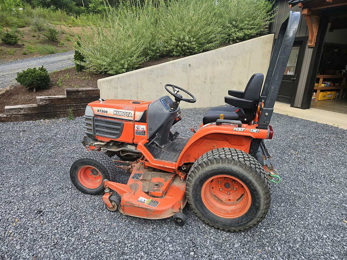 Kubota B7300 compact tractor with mower deck and roll bar, orange color, outdoor setting.