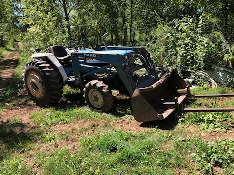 Ford 1920 compact utility tractor with front loader and cab, parked on grassy area near trees.