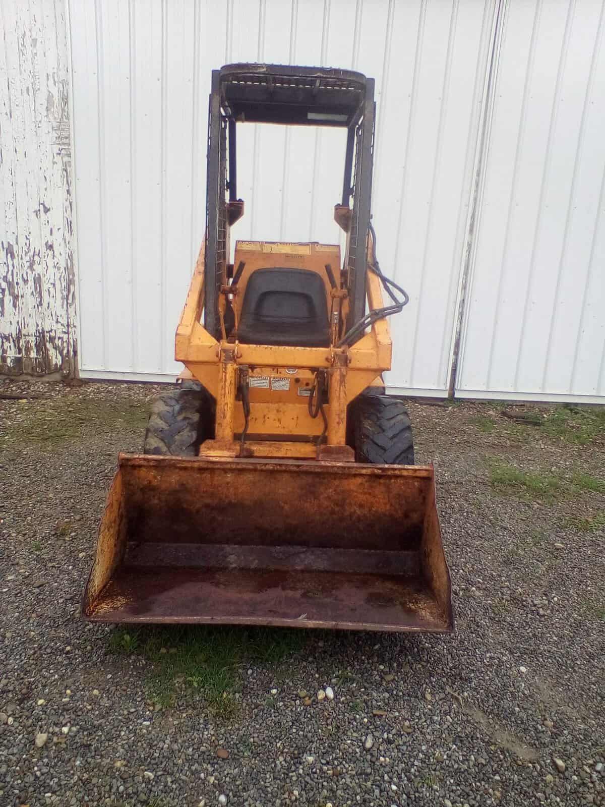 image000002 (10) Yellow skid steer loader with front bucket attachment in front of a white metal building.