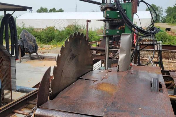 Rusty circular saw mill with hydraulic arm and control box on outdoor railway track.