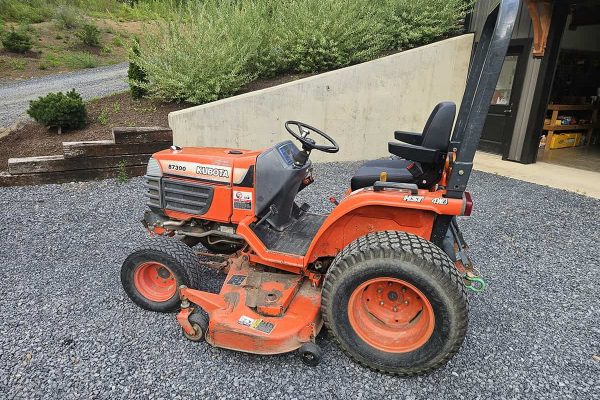 Kubota B7300 compact tractor with mower deck and roll bar, orange color, outdoor setting.