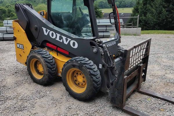 2019 Volvo MC85C skid steer loader with enclosed cab and fork attachment on gravel surface.
