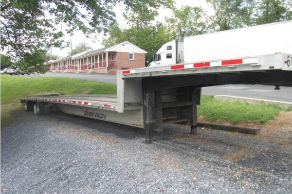Flatbed trailer with black and silver color parked on gravel surface near a residential building.