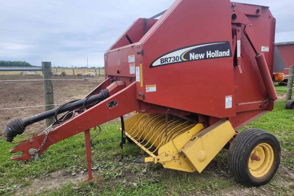 New Holland BR730 round baler for hay and straw collection, red color, with yellow pickup mechanism, on a farm field.